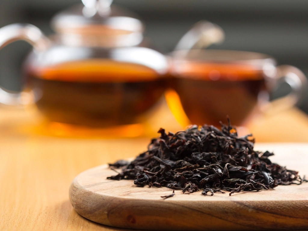 Pile of loose black tea leaves on a wooden board with a blurry teapot and cup behind