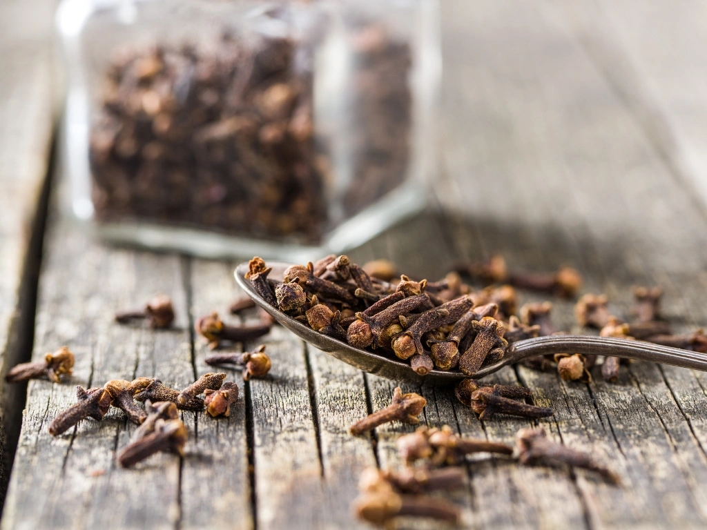 Spoonful of whole dried cloves on a rustic wooden table with a blurry spice jar in the background