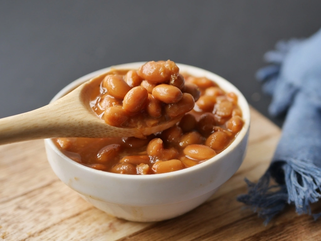 Wooden spoon scooping baked beans in tomato sauce from a white bowl