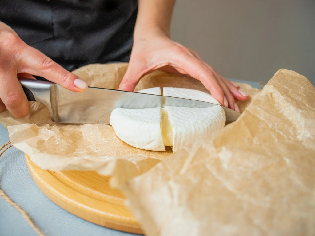 Person cutting a round of soft cheese into quarters with a knife