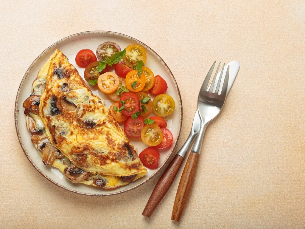 Plate of mushroom omelet with a side of tomatoes, served on a brown surface next to a fork and a knife