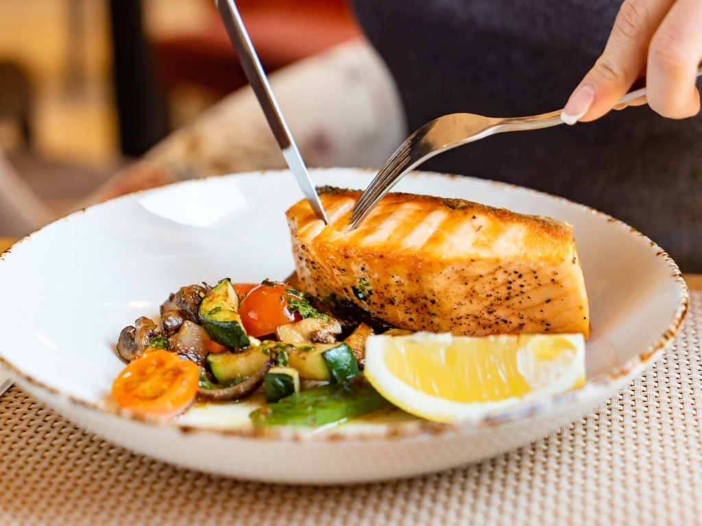 Person slicing salmon steak served in a bowl with vegetables and lemon