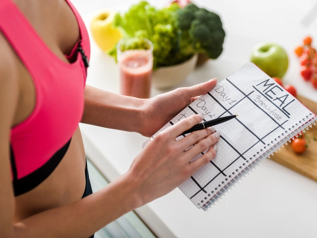 Woman in sportswear holding a notebook and a pen to create a meal plan, with various fruits and vegetables, a cutting board, and a glass of smoothie arranged on a white countertop