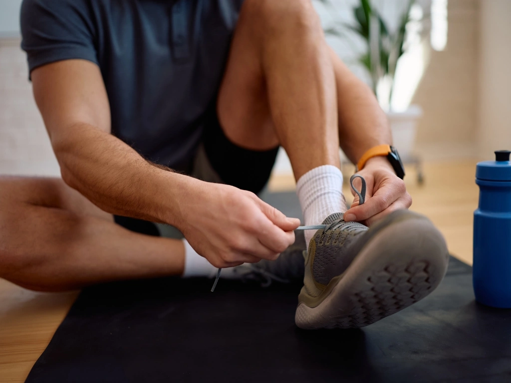 an tying his shoelace while sitting on a yoga mat next to a water bottle, getting ready to work out indoors