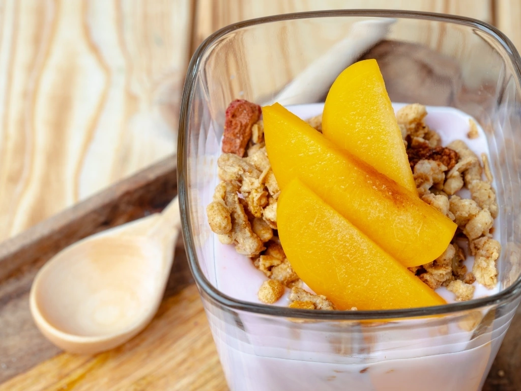 Yogurt parfait with peach slices and granola in a square-shaped glass, next to a wooden spoon