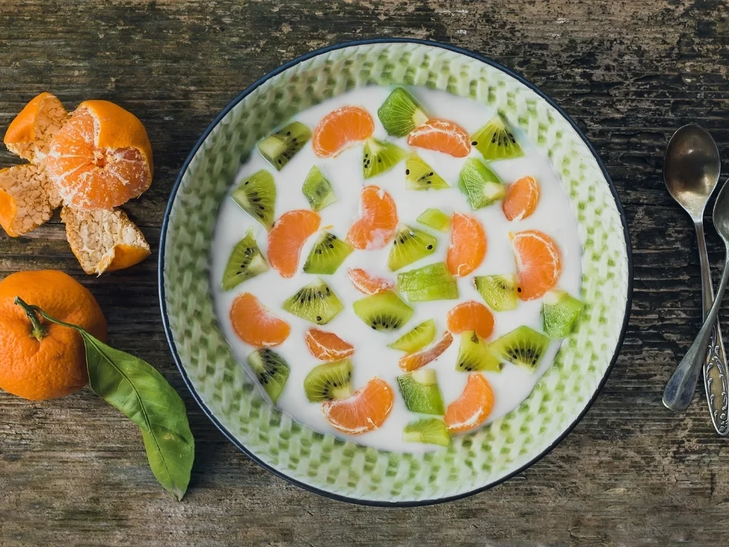 A bowl of sliced oranges and kiwi served with milk in a green bowl, next to spoons and a couple of oranges