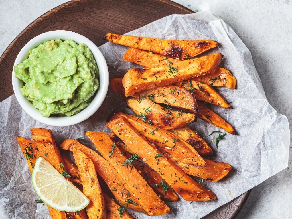 Roasted carrot sticks served on a wooden plate with guacamole and a slice of lemon