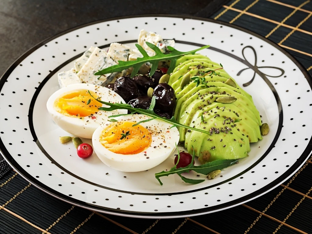 Plate of sliced avocado and boiled eggs served with cheese, olives, and herbs