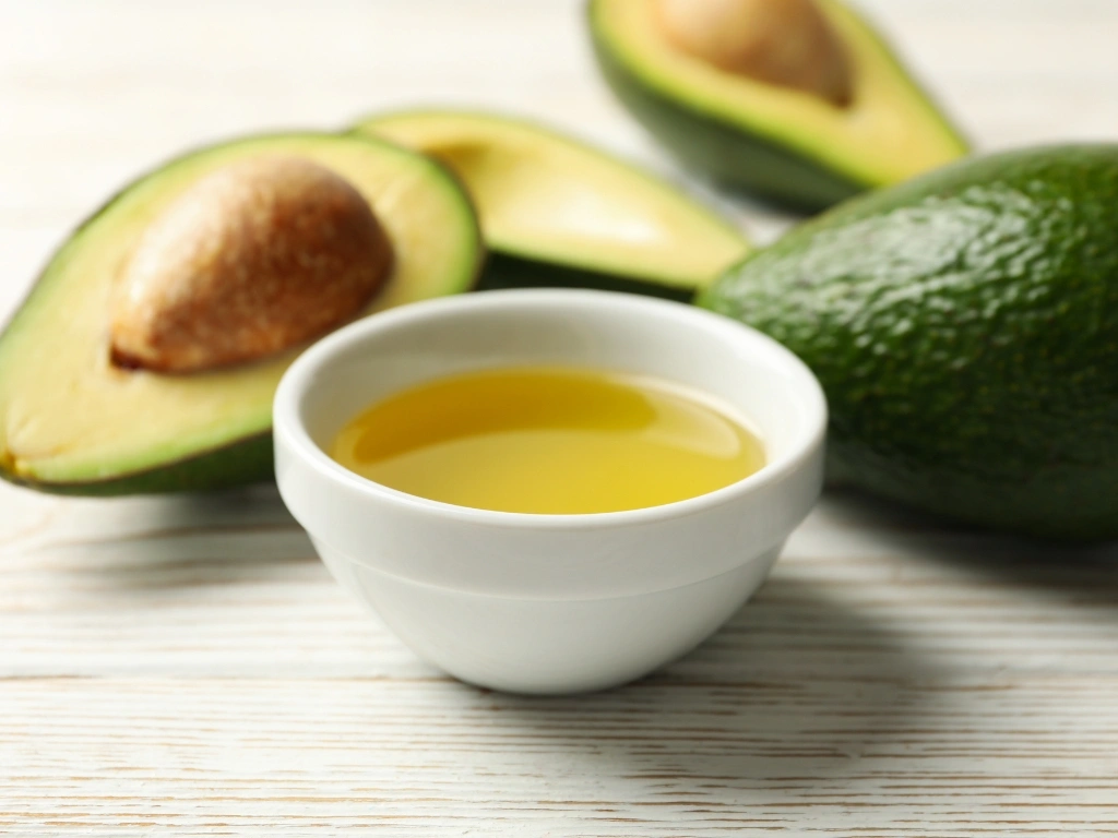 Small white bowl of avocado oil surrounded by whole and halved avocados