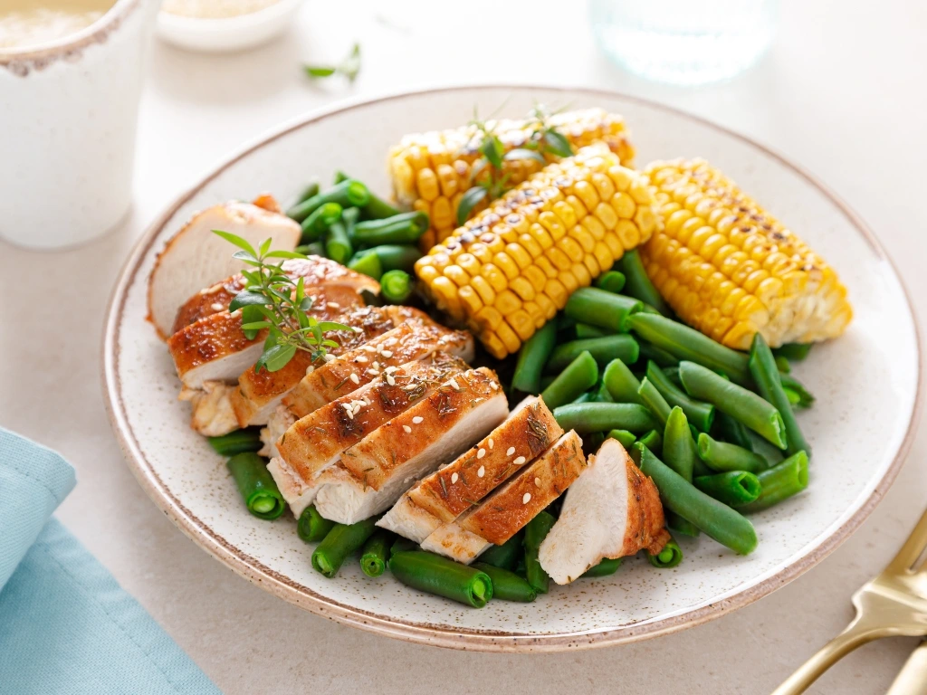 Slices of herb-crusted grilled chicken breast served with green beans and corn on the cob