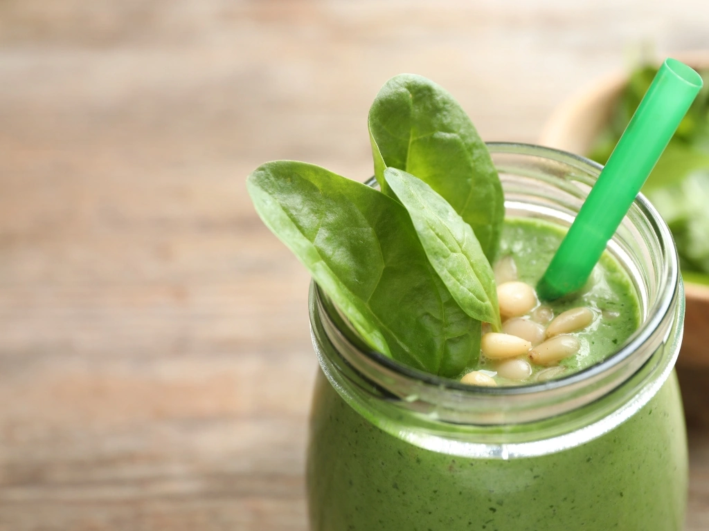 Spinach smoothie served in a glass jar with a straw, raw spinach leaves, and nuts