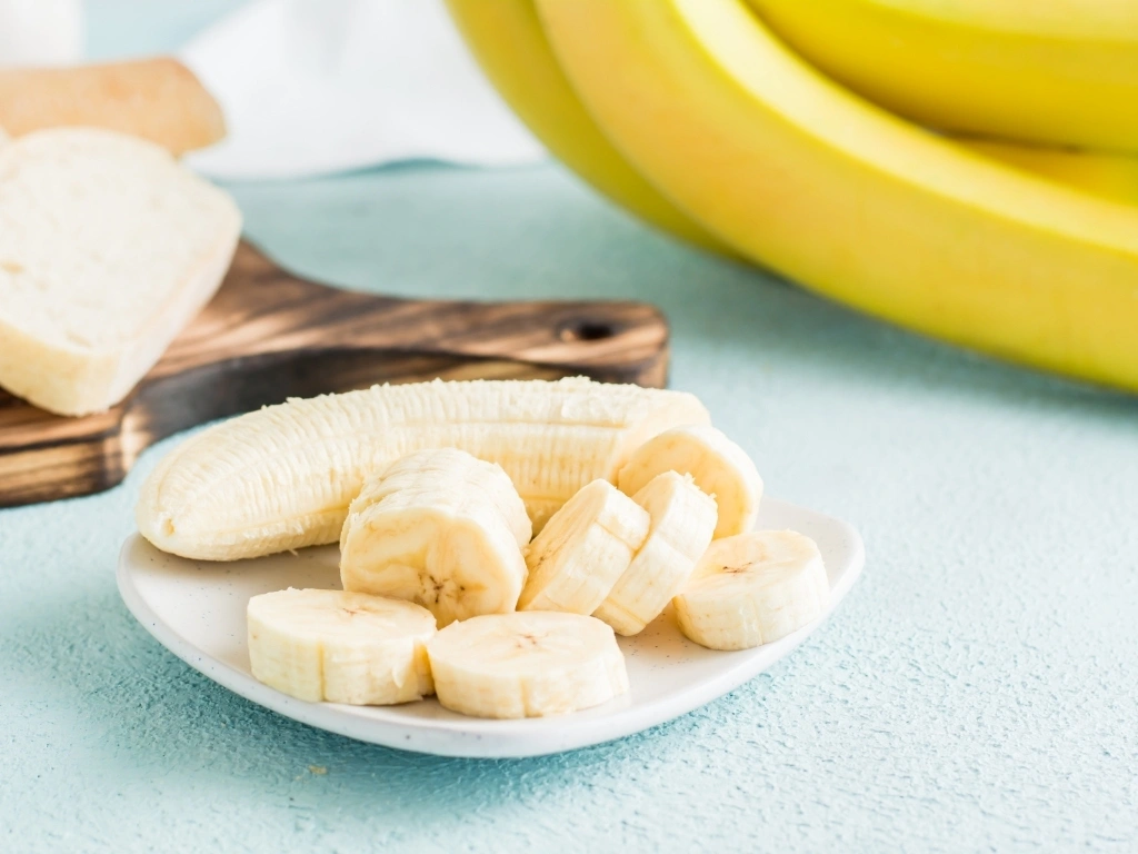 Sliced banana on a white plate with a bunch of unpeeled bananas behind it