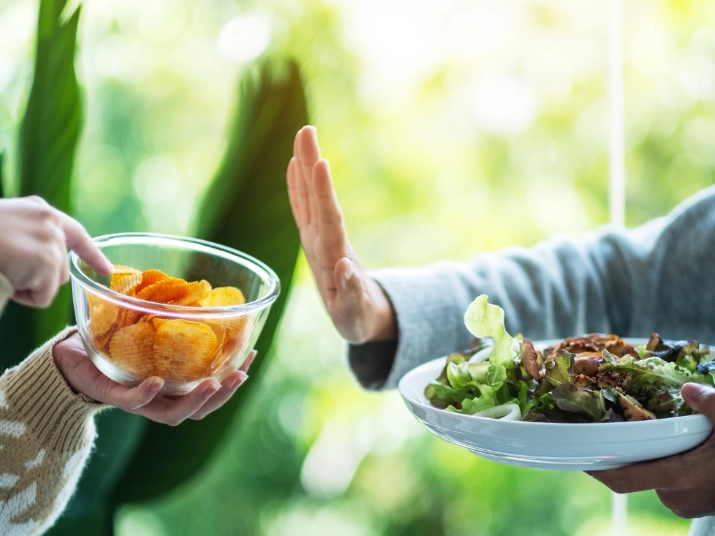 Person in a blue top holding a plate of salad while gesturing to decline a bowl of potato chips offered by another person in a brown top