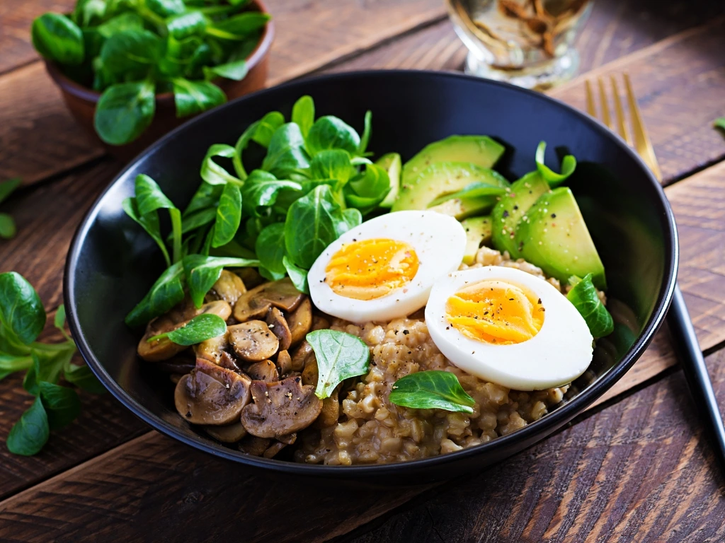 Oatmeal porridge with sliced avocado, mushrooms, leafy greens, and sliced egg in a black bowl, served with a fork and loose greens on a wooden table