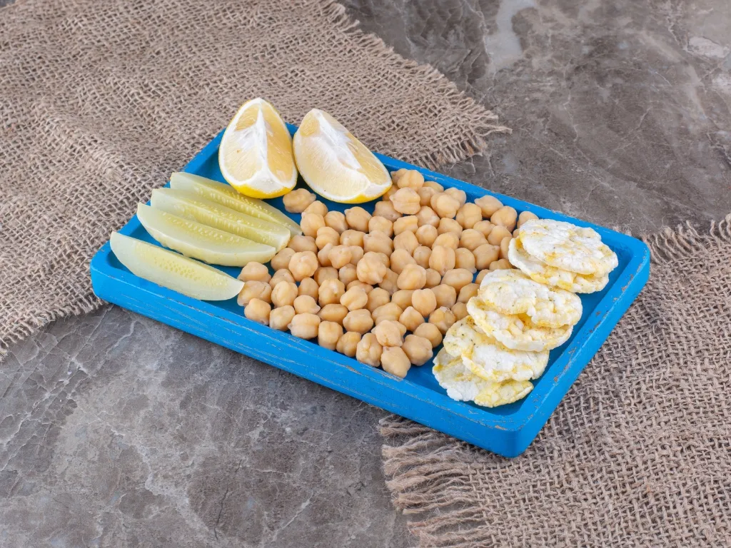 Chickpeas, sliced lemons, and white rice cakes presented on a blue wooden tray against a textured gray surface with a burlap cloth accent