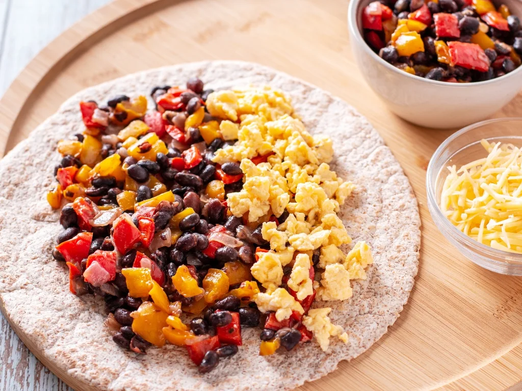 Tortilla topped with black beans, salsa, avocado, and an egg, served on a round wooden tray alongside a small bowl of shredded cheese and another bowl of the same toppings