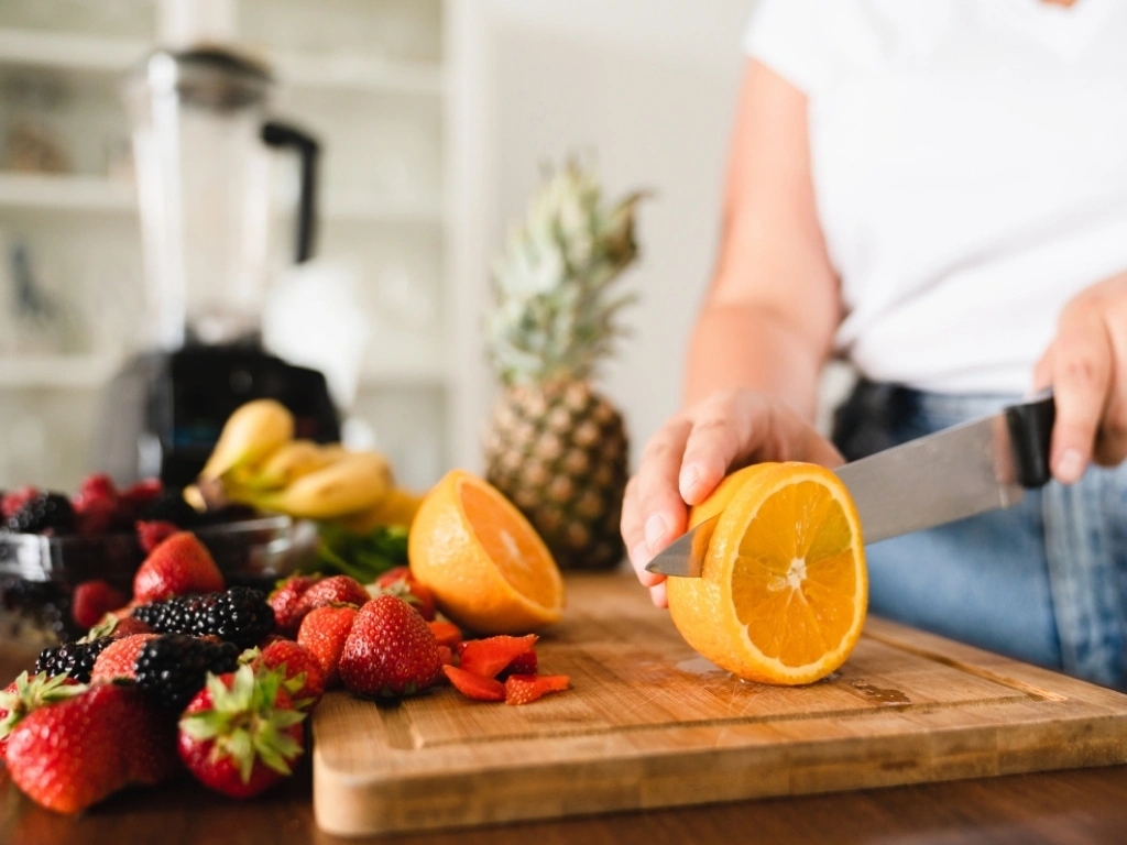 Person slicing oranges on a cutting board, with an assortment of other fruits across the table