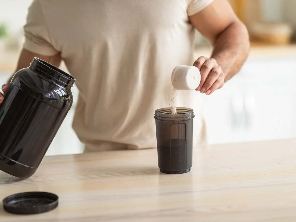 Person scooping protein powder from a large black container into a shaker bottle on a wooden table