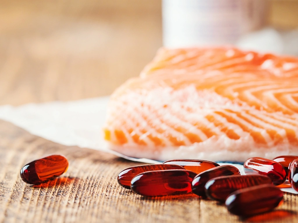 Red gel capsules on a wooden table with a fresh raw salmon fillet in the background