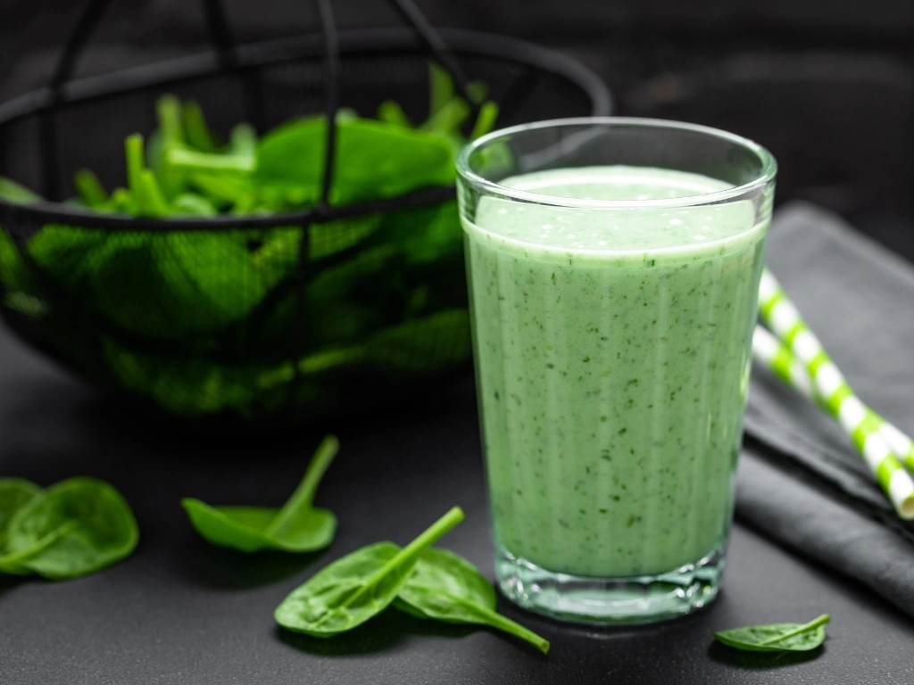 Glass of spinach smoothie, with straws and raw spinach in the background