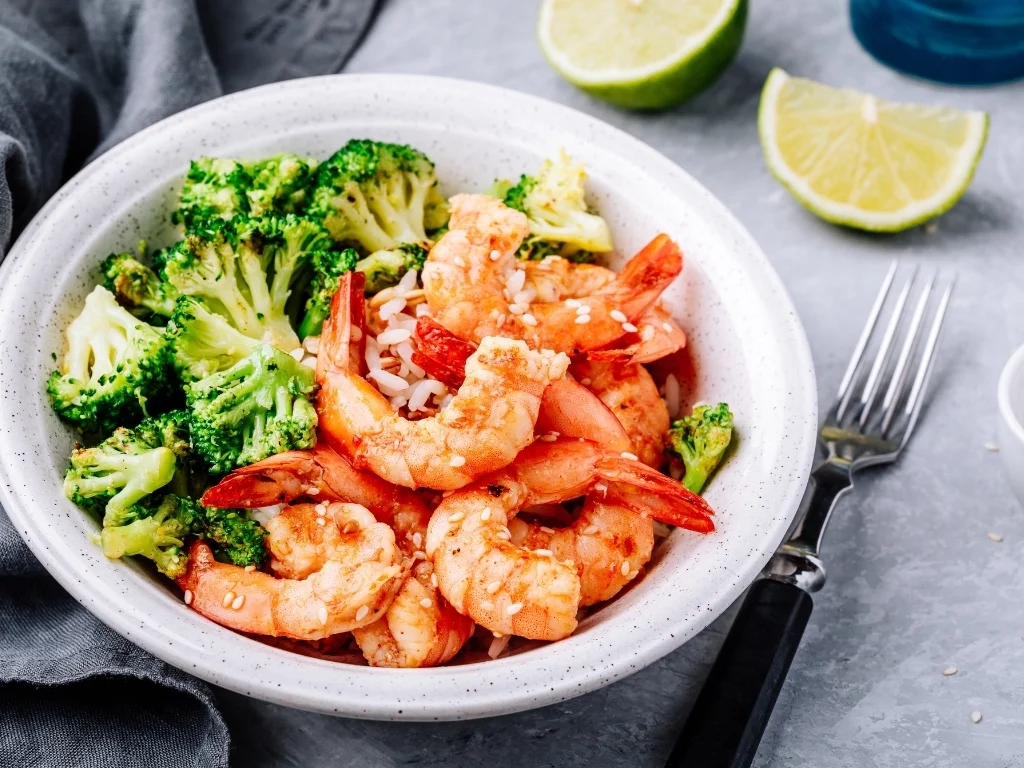 Bowl of grilled shrimp and broccoli on rice, next to a fork and lime wedges