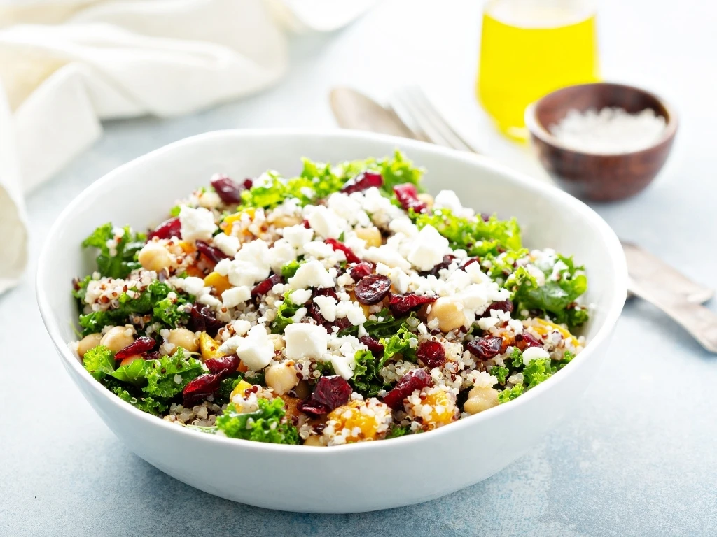 Bowl of quinoa salad with chickpeas, kale, and cottage cheese, with utensils and condiments in the background