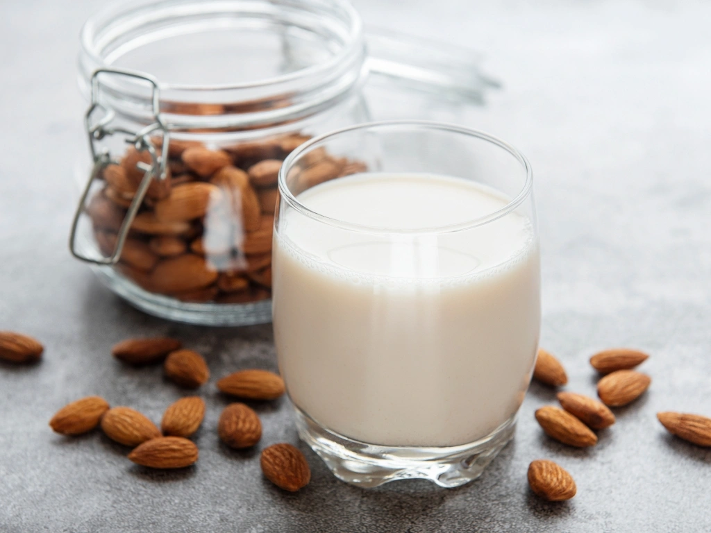 Glass of fresh almond milk next to a glass jar and scattered whole almonds on a countertop