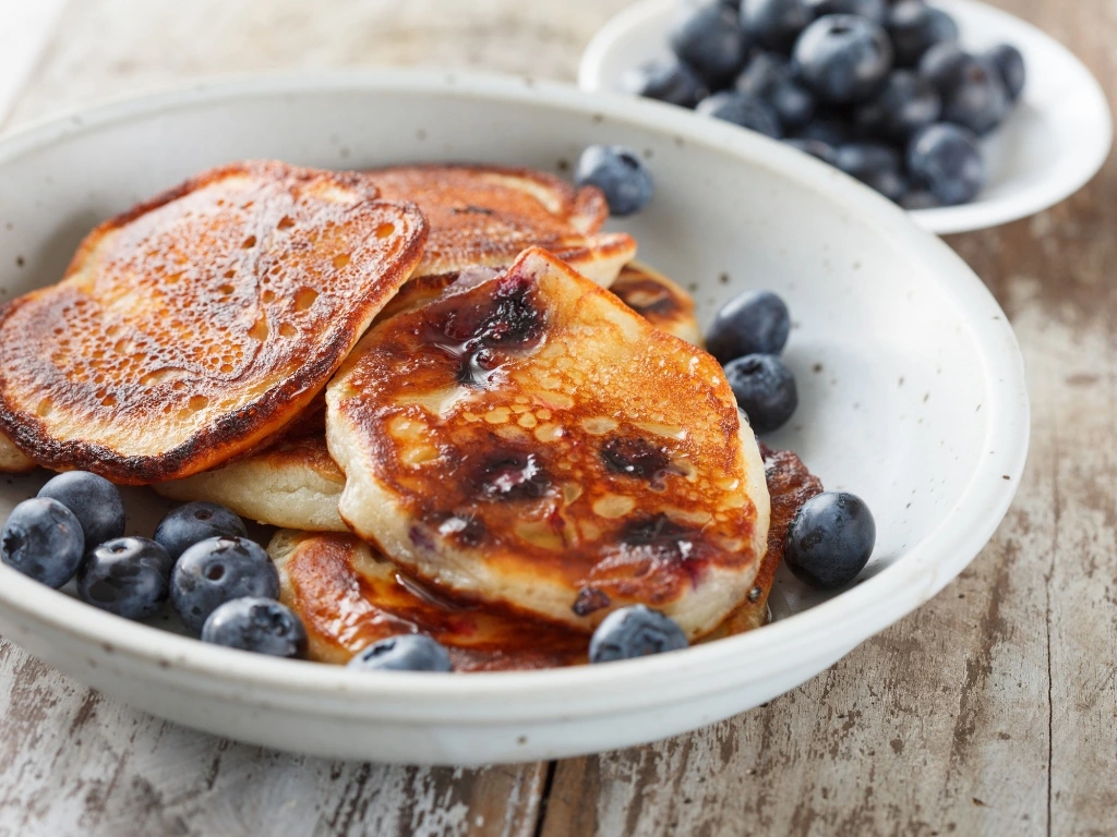 Bowl with homemade pancakes and fresh blueberries, next to a small dish with more berries