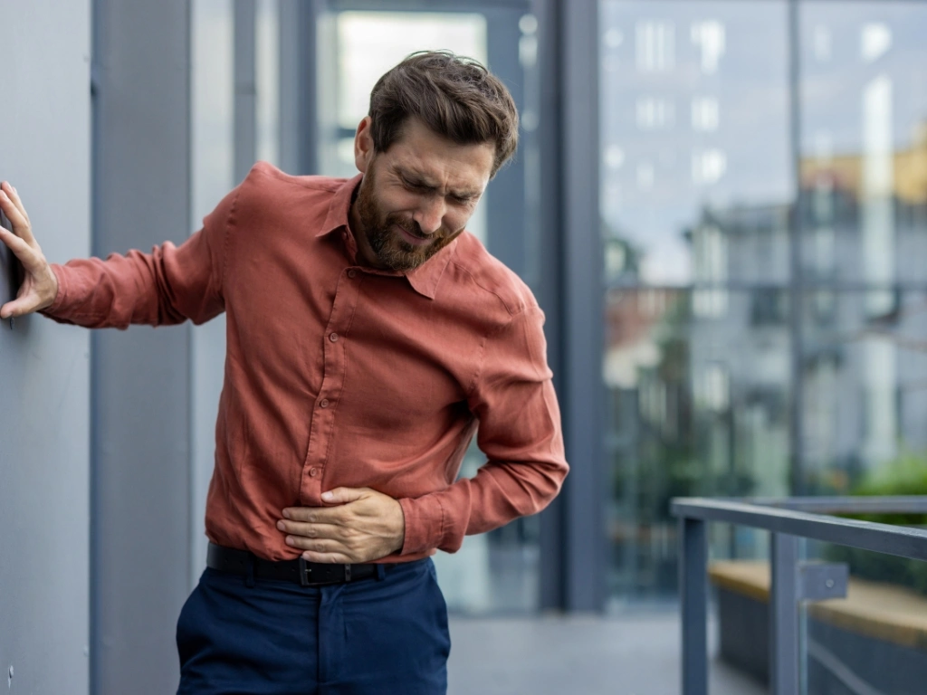 Man in a long-sleeved shirt leaning on a wall while holding lower abdomen in pain