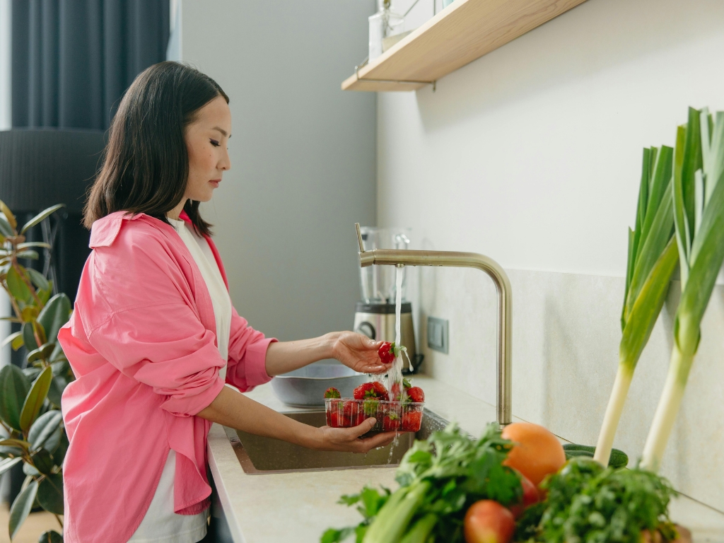 Woman in pink washing strawberries in the kitchen, with fresh produce on the countertop