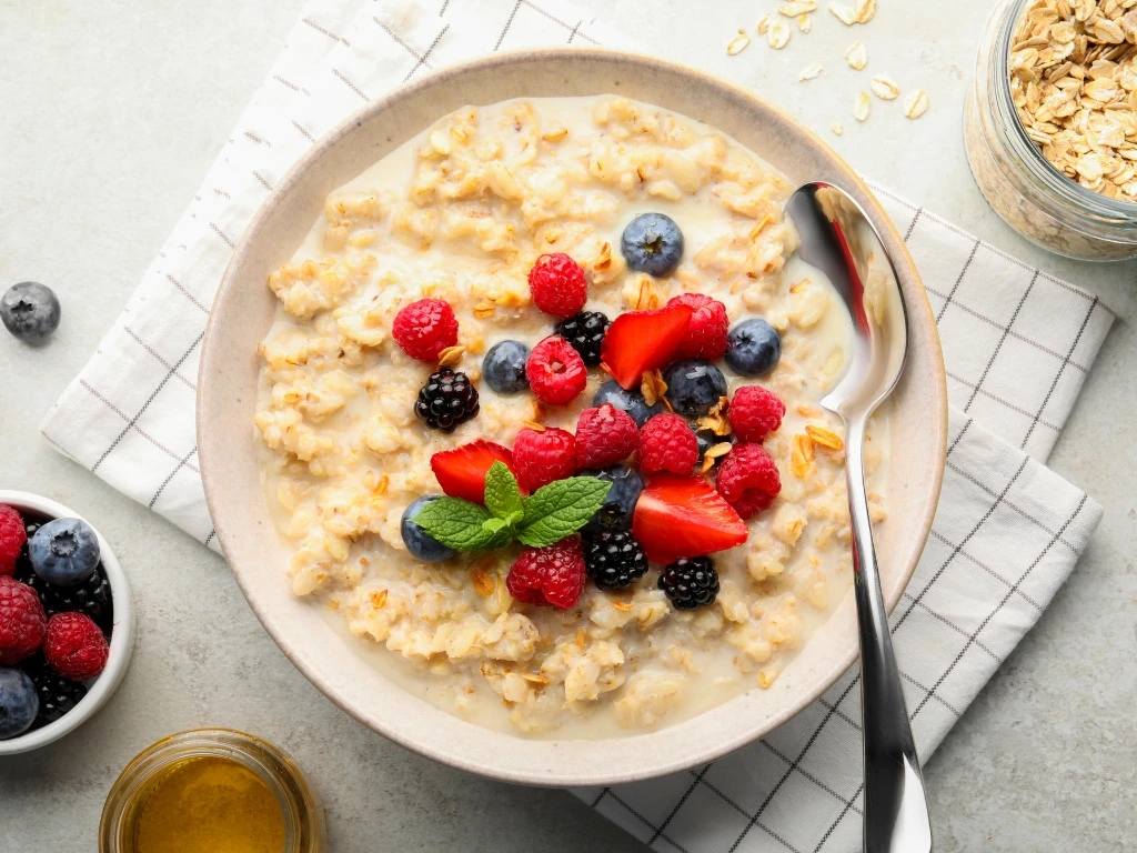 Bowl of oatmeal with different berries, served with a spoon on a tablecloth next to an open jar of uncooked oats, a small bowl of berries, and honey