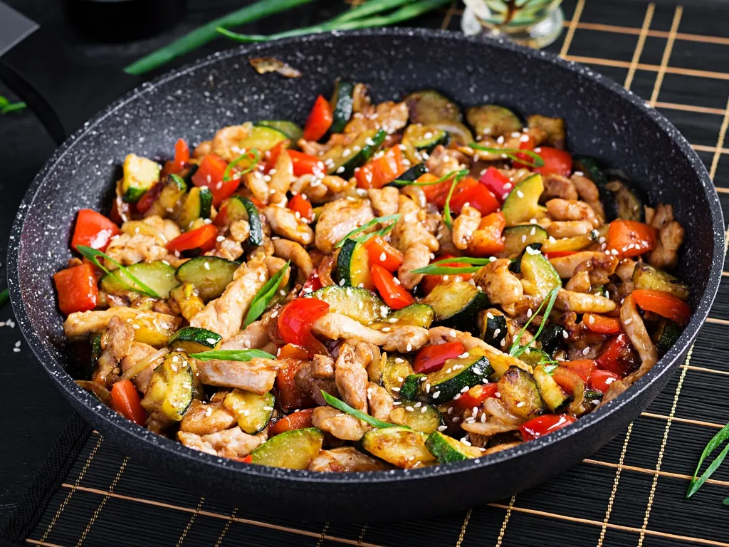 Stir-fried chicken with bell peppers and zucchini, garnished with sesame seeds and served on a black pan