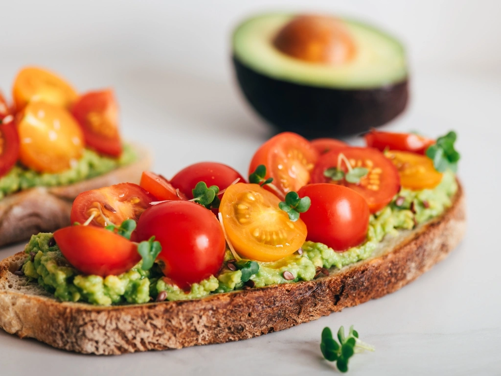 Two slices of whole-grain toast topped with mashed avocado and tomato slices, garnished with micro-greens and served next to a sliced avocado