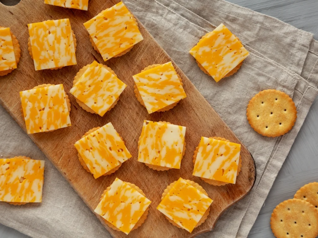 Various crackers topped with cheese squares scattered across a wooden board and a tablecloth