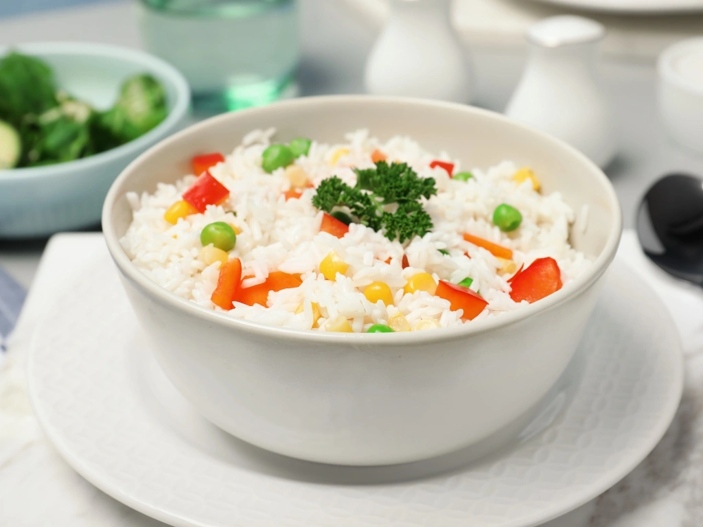 Bowl of plain rice and steamed vegetables on a plate next to a spoon, shakers, and a bowl of broccoli