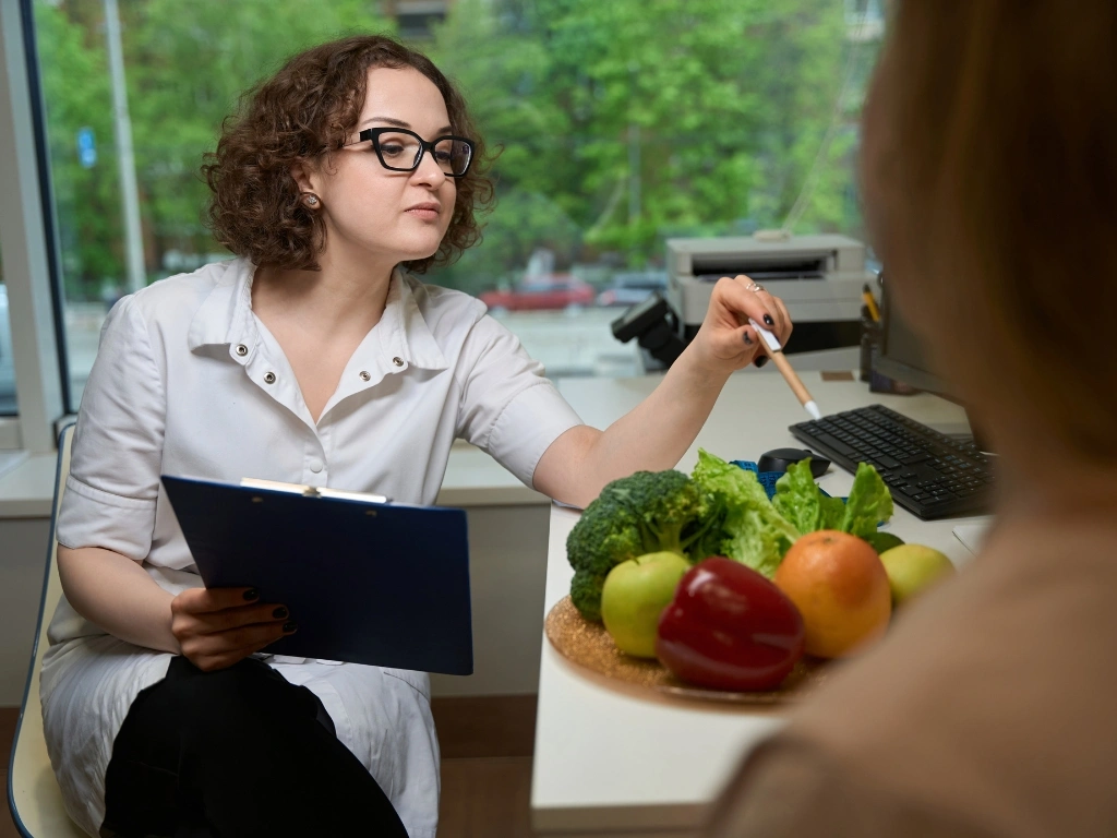 Dietitian during a medical nutrition therapy session. holding a pen in one hand and a clipboard in the other, pointing to a plate full of fruits and vegetables