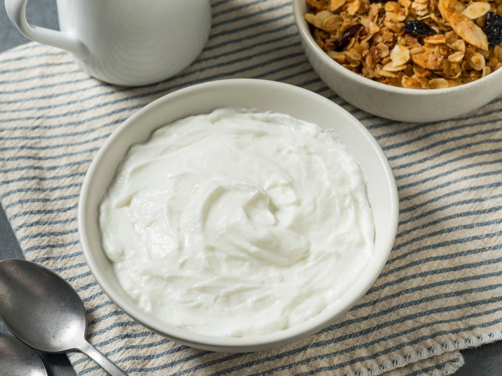 A bowl of plain yogurt surrounded by spoons, a mug, and a bowl of cereal