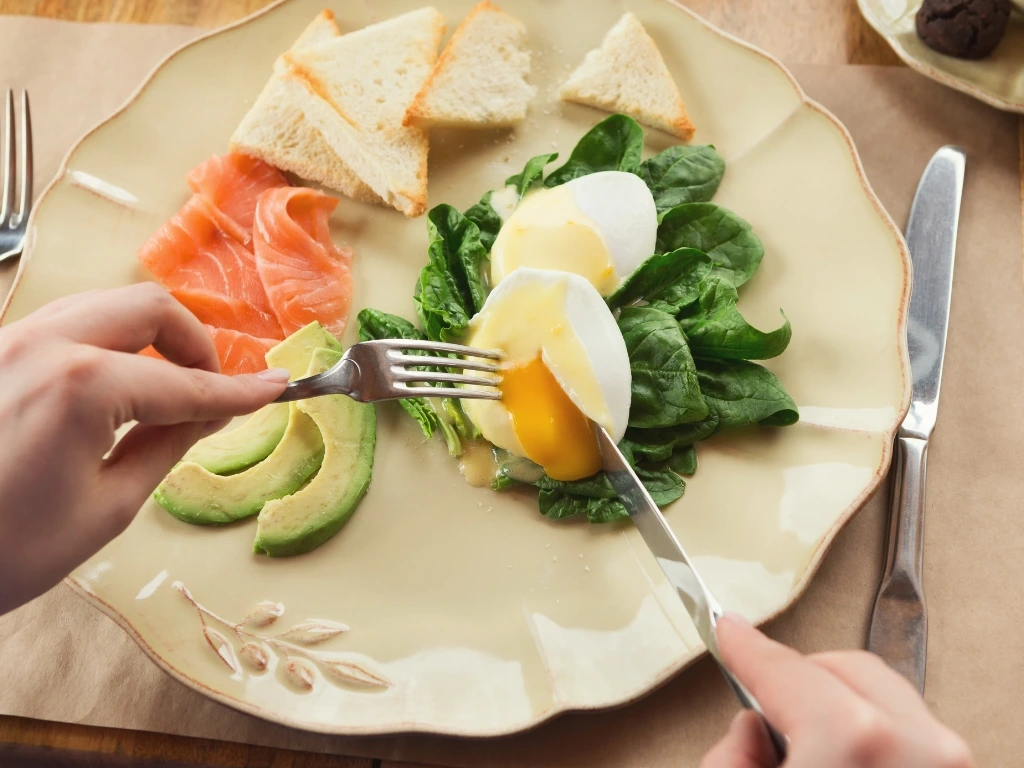 Person eating a small, balanced meal featuring bread, salmon, avocados, egg, and spinach