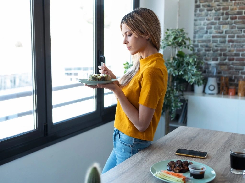 Woman in a yellow shirt eating a salad by a window in a modern kitchen in the daytime