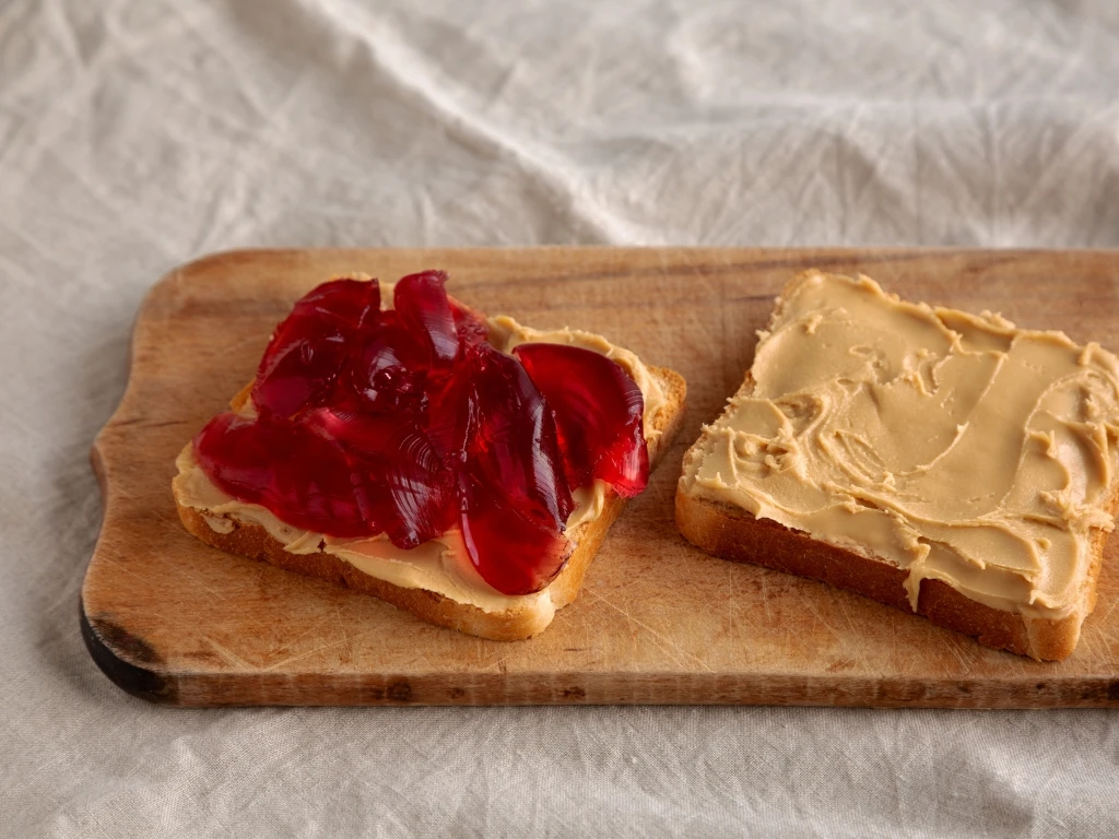 Two pieced of sliced bread, one topped with jelly and the other with peanut butter, both served on a wooden board