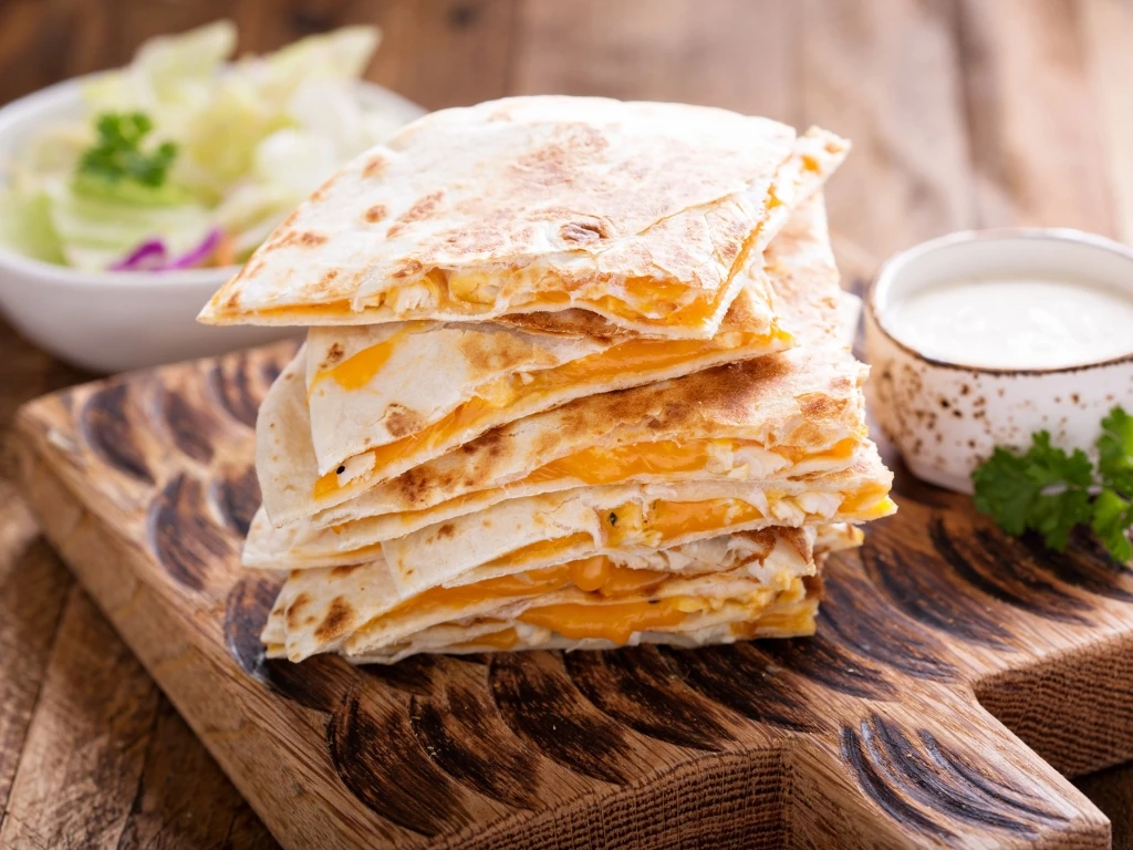Stack of cheesy quesadilla served on a wooden board with a white dip, next to a bowl of salad