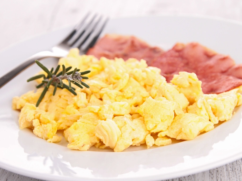 Plate of scrambled eggs served with fork, meat, and herbs