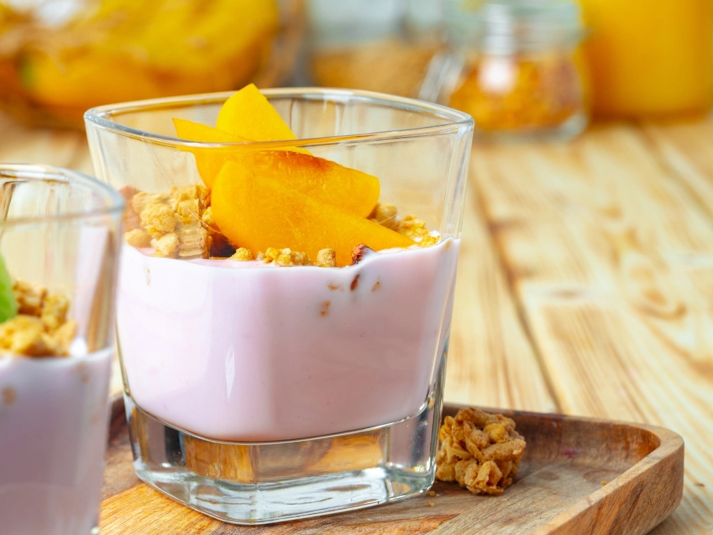 Yogurt with granola and fresh peach slices in a glass, served next to another yogurt cup with kiwi as the topping