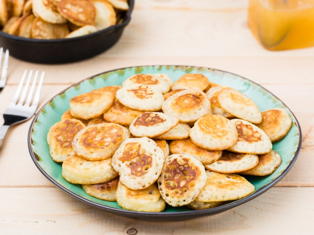 Mini pancakes in a blue-green plate, served next to two forks, a pan with more pancakes, and a jar of syrup