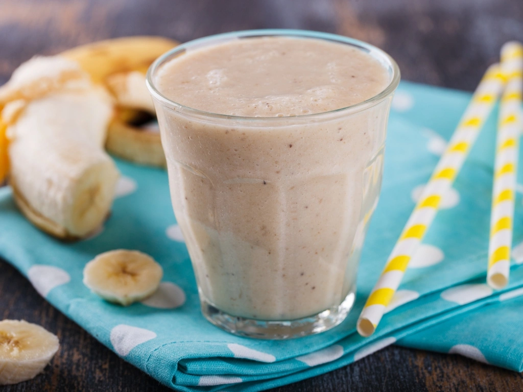 Clear glass filled with banana smoothie, served on a bright blue dotted tablecloth next to straws and sliced banana