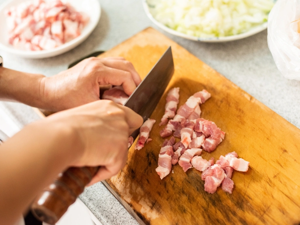 Person cutting raw pork into thin slices on a wooden board, with plates of meat and other ingredients across the countertop