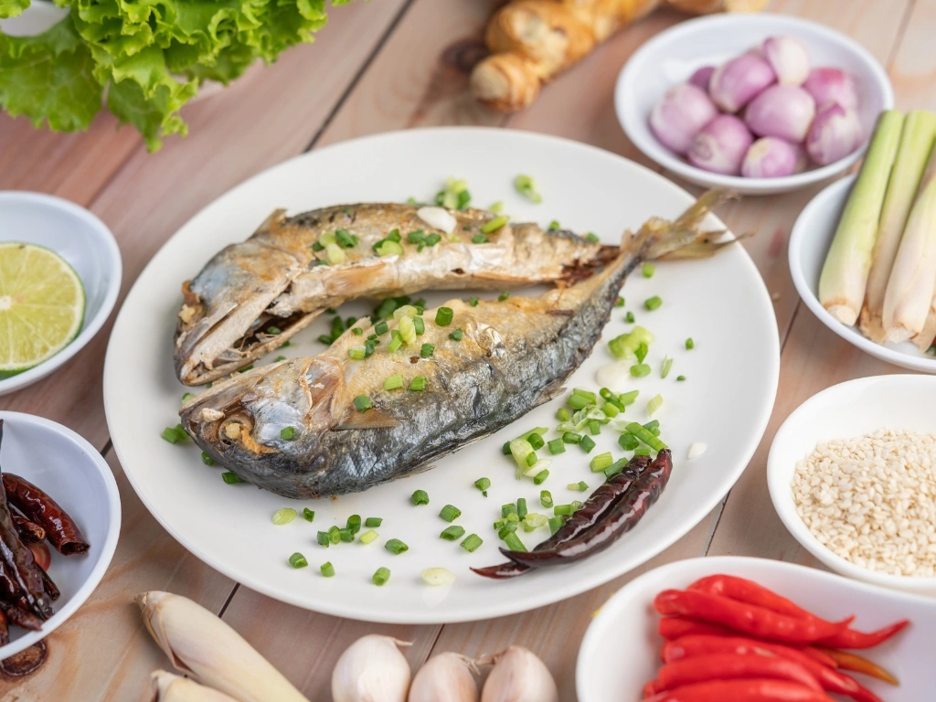 Two cooked mackerel garnished with green onion and served on a white dish next to vegetables and spices in smaller white bowls