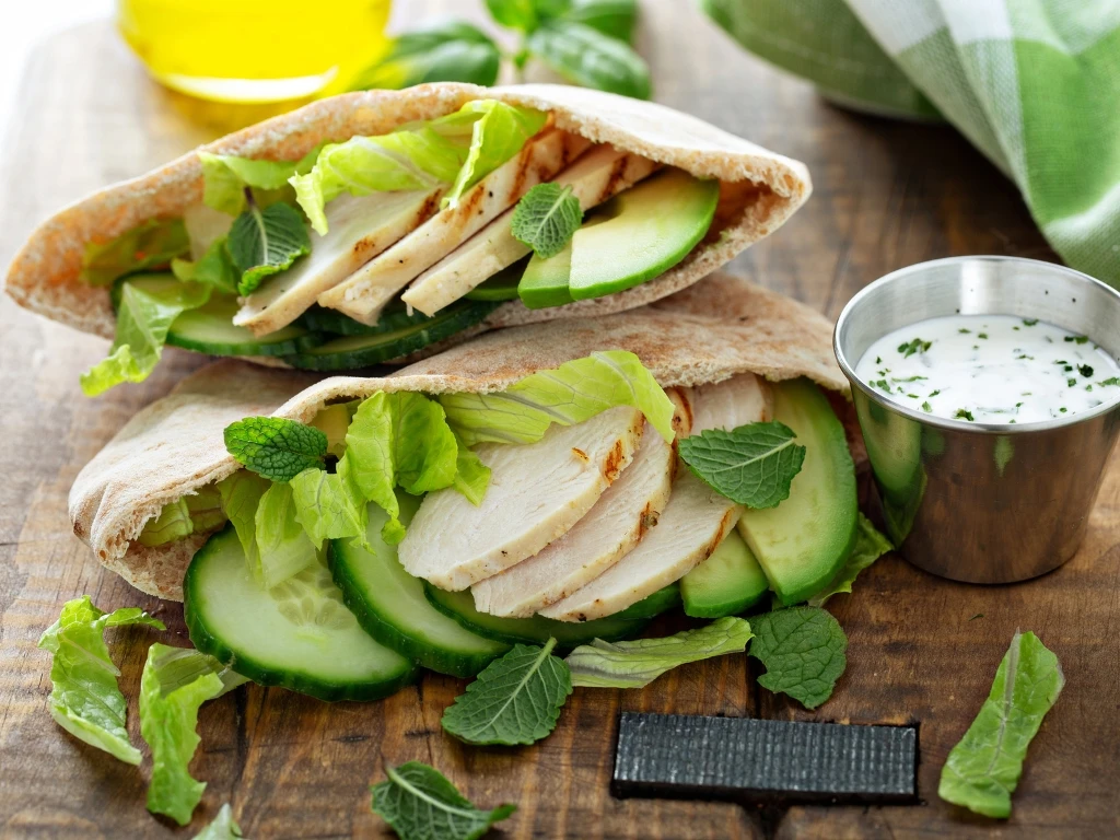 Two pita pockets filled with sliced grilled chicken, avocado, cucumber, and lettuce, served with cucumber tzatziki