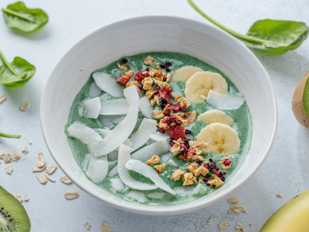 Green smoothie bowl topped with sliced bananas, coconut flakes, granola, and dried berries in a white bowl