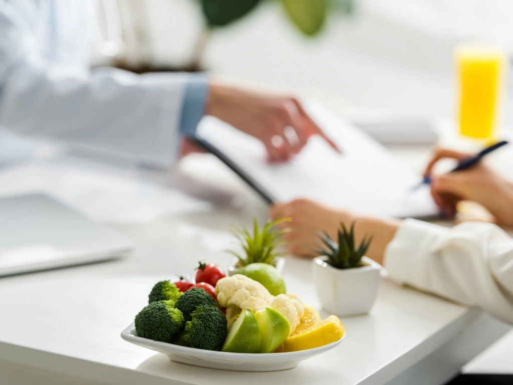A plate of different fruits and vegetables in the foreground, with a dietitian and patient discussing in the background